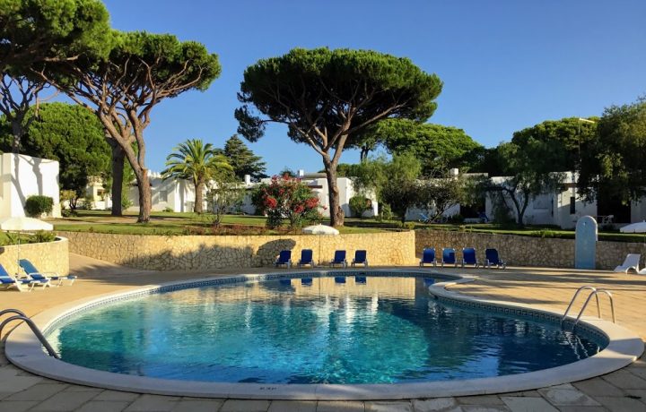 Prado Villas accommodation in Vilamoura Algarve Holiday external pool with pine trees surrounding pool area, with blue sun beds and umbrellas